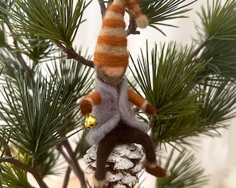 May include: A felt gnome ornament with a striped orange and white hat, sitting on a pine cone. The gnome is wearing a gray coat and brown pants and is holding a small gold bell.