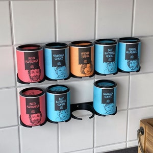 Könnte beinhalten: Ein Satz von acht Gewürzgläsern mit schwarzen Deckeln und bunten Etiketten. Die Etiketten zeigen Illustrationen von Personen und Text in deutscher Sprache. Die Gläser sind an einem schwarzen Metallgestell befestigt.