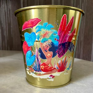 May include: A gold metal planter featuring a colourful illustration of a woman in a tropical setting. The woman is wearing a flower crown and a red skirt. The illustration includes palm leaves, flowers, and other tropical plants.