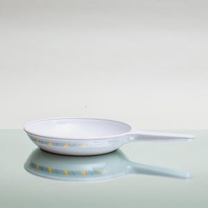 Puede incluir: Una pequeña sartén blanca con un mango largo. La sartén tiene un cuenco poco profundo con una banda decorativa de formas amarillas y azules alrededor del borde. La sartén está sobre una superficie reflectante.