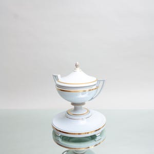 May include: A white and light blue porcelain lidded dish with gold trim. The dish has a pedestal base and a small finial on the lid. The design is symmetrical and elegant, suitable for serving or display.