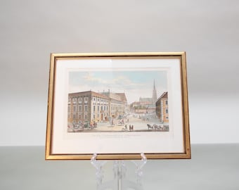 Puede incluir: Una impresión enmarcada de una ilustración vintage que representa una escena de calle de la ciudad con un gran edificio en primer plano y un campanario de iglesia en el fondo. La impresión está en un marco dorado y sobre un fondo blanco.