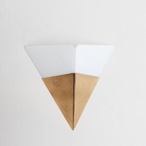May include: A contemporary wall light with a geometric design. The light has a triangular shape with a gold-coloured base and white, triangular top. The fixture is designed for wall mounting and provides ambient lighting.