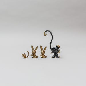 Könnte beinhalten: Eine Sammlung kleiner Metalltierfiguren, darunter drei Hasen und eine Maus. Die Hasenfiguren sind goldfarben, während die Maus und das letzte Tier schwarz sind. Die Maus hat einen langen, gebogenen Schwanz. Auf schlichtem Hintergrund.