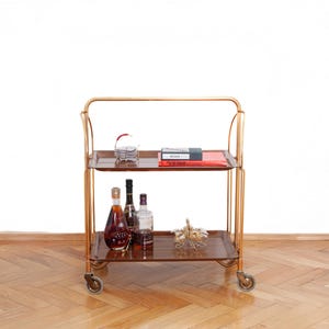 Carrito de bar con decoración de mediados de siglo: carrito de té plegable estilo Dinett de los años 60 con bandejas y estructura de metal dorado.