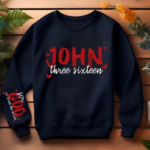 May include: Navy blue sweatshirt with red lettering that says "JOHN three sixteen" and red hearts. The left sleeve has red lettering that says "for GOD so loved the world" with red hearts.