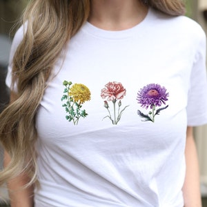 Op de afbeelding: Wit T-shirt met een bloemenprint met vier bloemen: een gele chrysant, een roze anjer, een paarse aster en een groen blad.