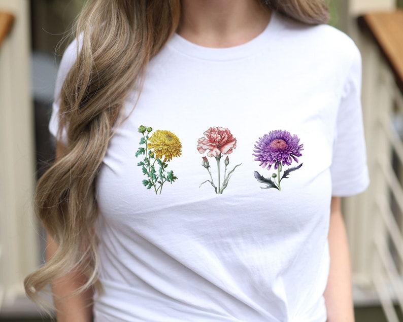 Op de afbeelding: Wit T-shirt met een bloemenprint met vier bloemen: een gele chrysant, een roze anjer, een paarse aster en een groen blad.