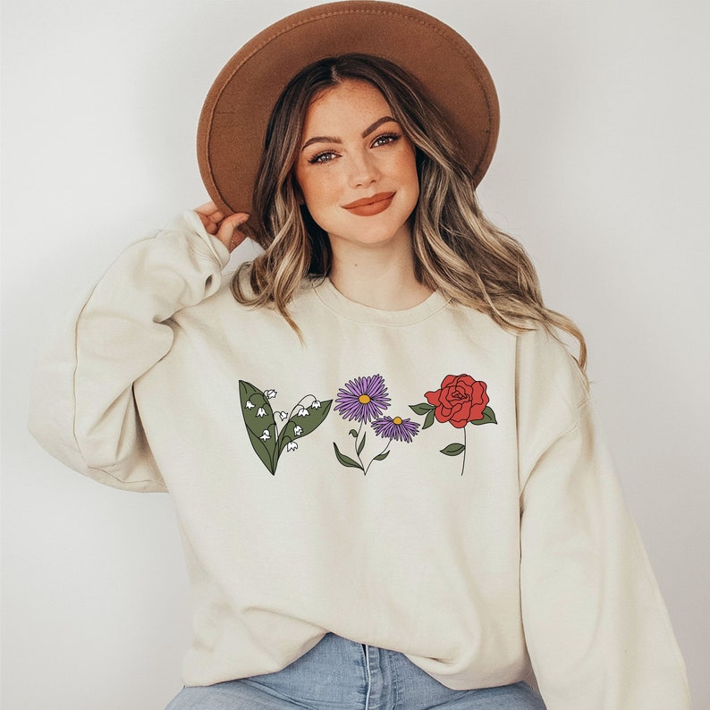 May include: A cream-colored sweatshirt with a floral design featuring a red rose, purple daisy, and white lily of the valley. The sweatshirt has a crew neck and long sleeves.