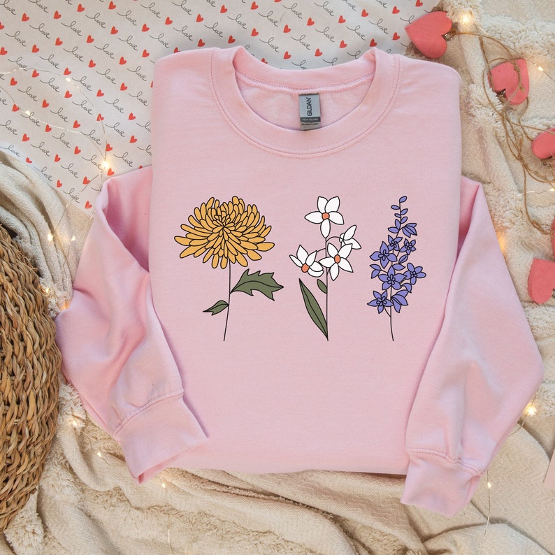 May include: A pink sweatshirt with a graphic of three stylized flowers. The flowers are yellow, white, and purple.