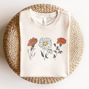 May include: A white t-shirt with a simple line drawing of three flowers. The flowers are a daisy, a carnation, and a carnation. The flowers are in shades of orange, white, and green.