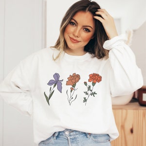 May include: White sweatshirt with a floral design featuring an iris, a carnation, and a marigold. The flowers are outlined in black and filled with shades of purple, orange, and red.