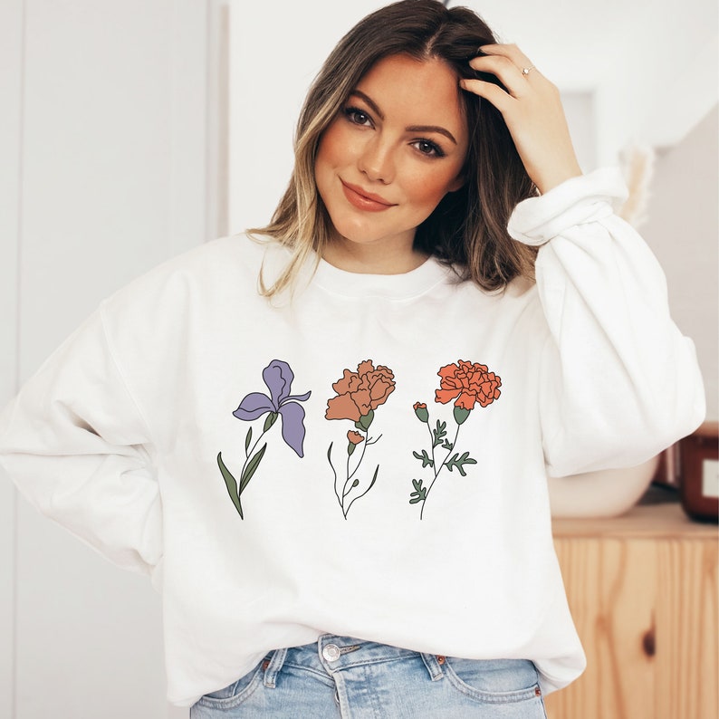 May include: White sweatshirt with a floral design featuring an iris, a carnation, and a marigold. The flowers are outlined in black and filled with shades of purple, orange, and red.