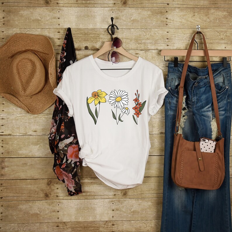 May include: White t-shirt with a floral design featuring a daisy, daffodil, and other flowers. The shirt is hanging on a wooden hanger with a straw hat and a denim skirt in the background.