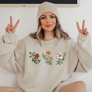Op de afbeelding: Een beige sweatshirt met een bloemenprint met een rode roos, een gele bloem en een witte lelie.