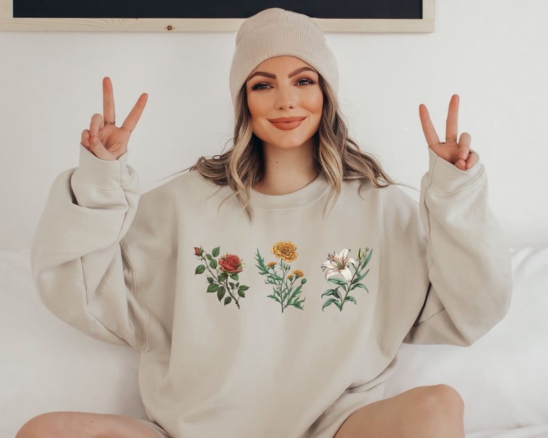 Op de afbeelding: Een beige sweatshirt met een bloemenprint met een rode roos, een gele bloem en een witte lelie.