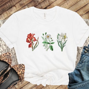 Op de afbeelding: Wit T-shirt met een bloemenprint met drie verschillende bloemen: een rode iris, een witte madeliefje en een witte narcis.