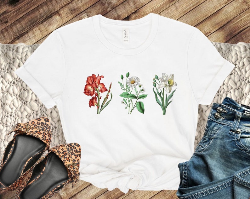 Op de afbeelding: Wit T-shirt met een bloemenprint met drie verschillende bloemen: een rode iris, een witte madeliefje en een witte narcis.