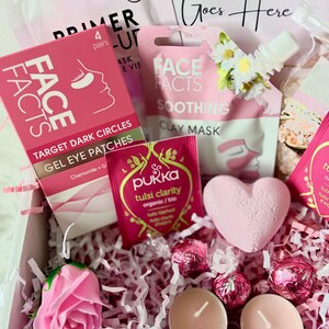 May include: Pink and white gift box filled with self-care items, including face masks, bath bombs, tea, and chocolates. The box is decorated with pink tissue paper and a pink rose.
