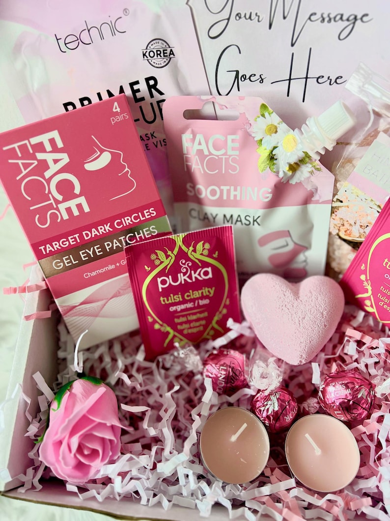 May include: Pink and white gift box filled with self-care items, including face masks, bath bombs, tea, and chocolates. The box is decorated with pink tissue paper and a pink rose.