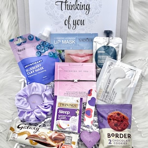 May include: A gift box containing self-care products. Includes a blueberry clay mask, lip mask, eye mask, scrunchie, bracelet, tea, chocolate, and cookies. The box is labelled with the words "Thinking of you."