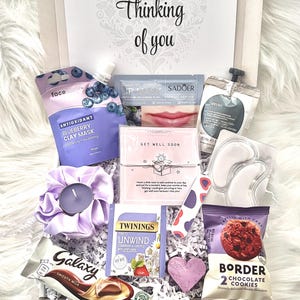 May include: A gift box filled with self-care items. Includes a face mask, lip mask, eye patches, tea, chocolate, cookies, a candle, and a heart-shaped decoration. The box is decorated with the words "Thinking of you".