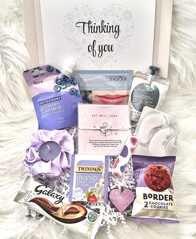 May include: A gift box filled with self-care items. Includes a face mask, lip mask, eye patches, tea, chocolate, cookies, a candle, and a heart-shaped decoration. The box is decorated with the words "Thinking of you".