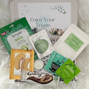 May include: A gift box filled with self-care items, including a face mask, eye masks, bath salts, tea, chocolate, and a candle. The box is decorated with green and white shredded paper and has a label that says "Enjoy Your Treats".