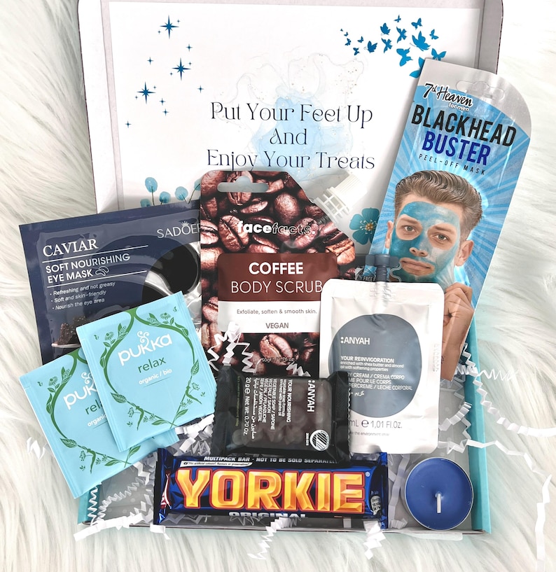 May include: A gift box filled with self-care items, including a coffee body scrub, a blackhead peel-off mask, a blue tea light candle, a chocolate bar, and several small packets of bath and beauty products. The box is decorated with a blue and white patterned paper and the text "Put Your Feet Up And Enjoy Your Treats".
