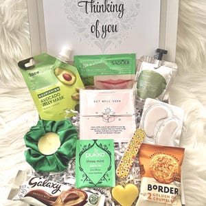 May include: A gift box filled with self-care items, including an avocado face mask, lip balm, eye patches, a scrunchie, tea, chocolate, a nail file, and a heart-shaped soap. The box is decorated with the words "Thinking of you."