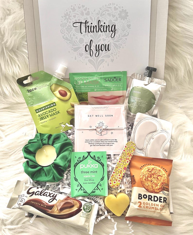 May include: A gift box filled with self-care items, including an avocado face mask, lip balm, eye patches, a scrunchie, tea, chocolate, a nail file, and a heart-shaped soap. The box is decorated with the words "Thinking of you."
