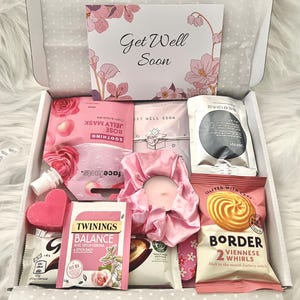 May include: A gift box filled with self-care items. The box contains a face mask, tea, a candle, a scrunchie, and cookies. The box has a card that says "Get Well Soon" with floral accents. The items are primarily pink and white.
