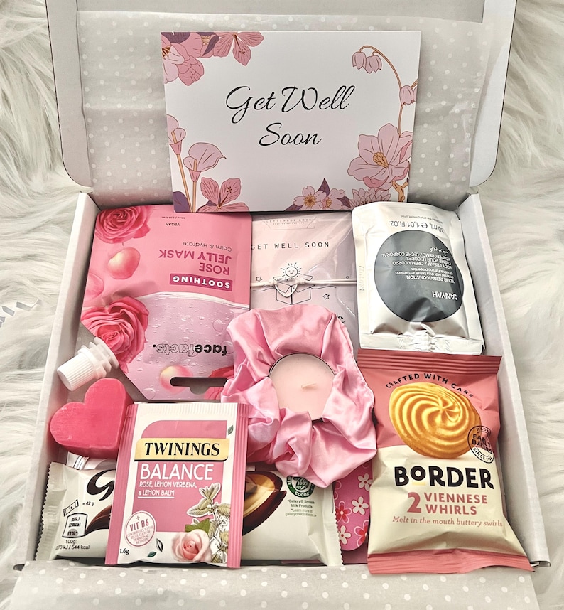 May include: A gift box filled with self-care items. The box contains a face mask, tea, a candle, a scrunchie, and cookies. The box has a card that says "Get Well Soon" with floral accents. The items are primarily pink and white.