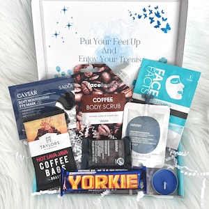 May include: A gift box filled with self-care items, including a coffee body scrub, face mask, eye mask, and a chocolate bar. The box is decorated with a blue and white watercolor background and the text "Put Your Feet Up And Enjoy Your Treats".
