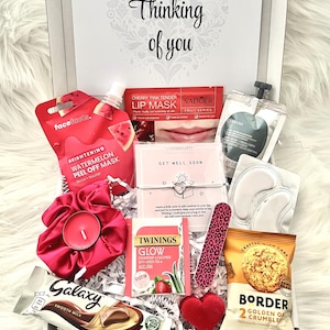 May include: A gift box filled with self-care items. Includes a face mask, lip mask, tea, chocolate, snacks, a candle, and a nail file. The box is white with the words "Thinking of you" on the lid.