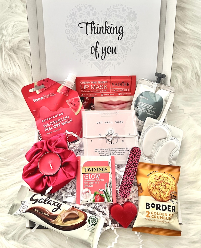 May include: A gift box filled with self-care items. Includes a face mask, lip mask, tea, chocolate, snacks, a candle, and a nail file. The box is white with the words "Thinking of you" on the lid.