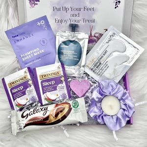 May include: A gift box filled with self-care items. Includes a face mask, eye mask, tea bags, a chocolate bar, a heart-shaped soap, and a lavender scrunchie with a candle. The box has the text "Put Up Your Feet and Enjoy Your Treat".