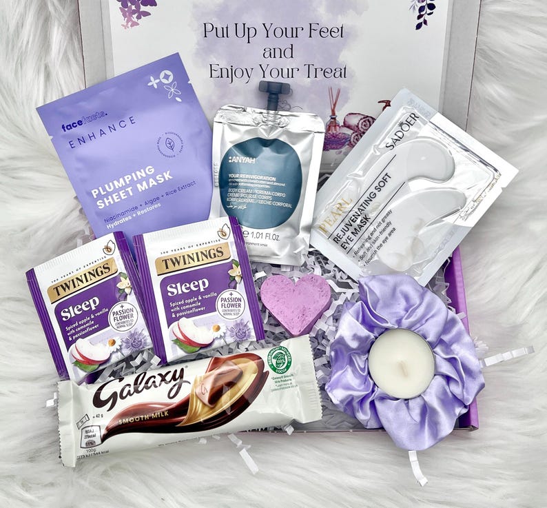 May include: A gift box filled with self-care items. Includes a face mask, eye mask, tea bags, a chocolate bar, a heart-shaped soap, and a lavender scrunchie with a candle. The box has the text "Put Up Your Feet and Enjoy Your Treat".