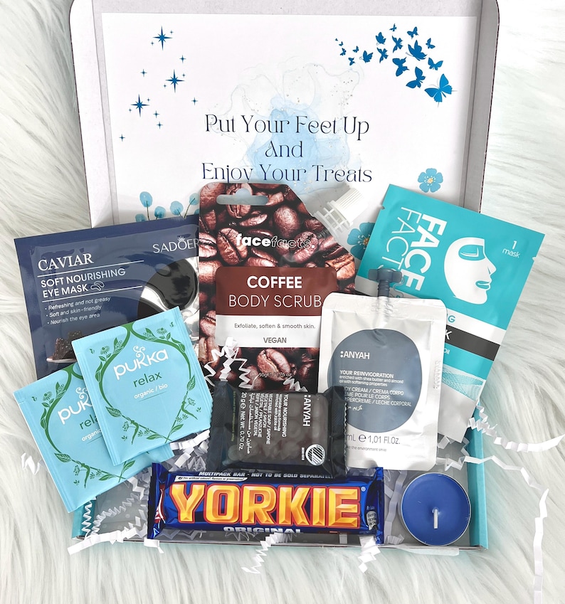 May include: A gift box filled with self-care items, including a coffee body scrub, face mask, eye mask, tea bags, a chocolate bar, and a candle. The box is decorated with a blue and white watercolor design and the text "Put Your Feet Up And Enjoy Your Treats".