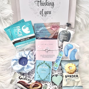 May include: A gift box with the text "Thinking of you" contains beauty products and treats. Items include face masks, lip mask, eye gel pads, a candle, tea, chocolate, shortbread cookies, and a heart-shaped decoration. The box is filled with white crinkle paper.