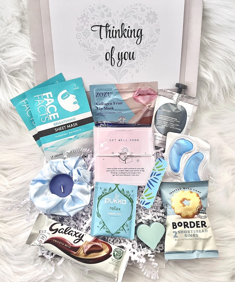 May include: A gift box with the text "Thinking of you" contains beauty products and treats. Items include face masks, lip mask, eye gel pads, a candle, tea, chocolate, shortbread cookies, and a heart-shaped decoration. The box is filled with white crinkle paper.