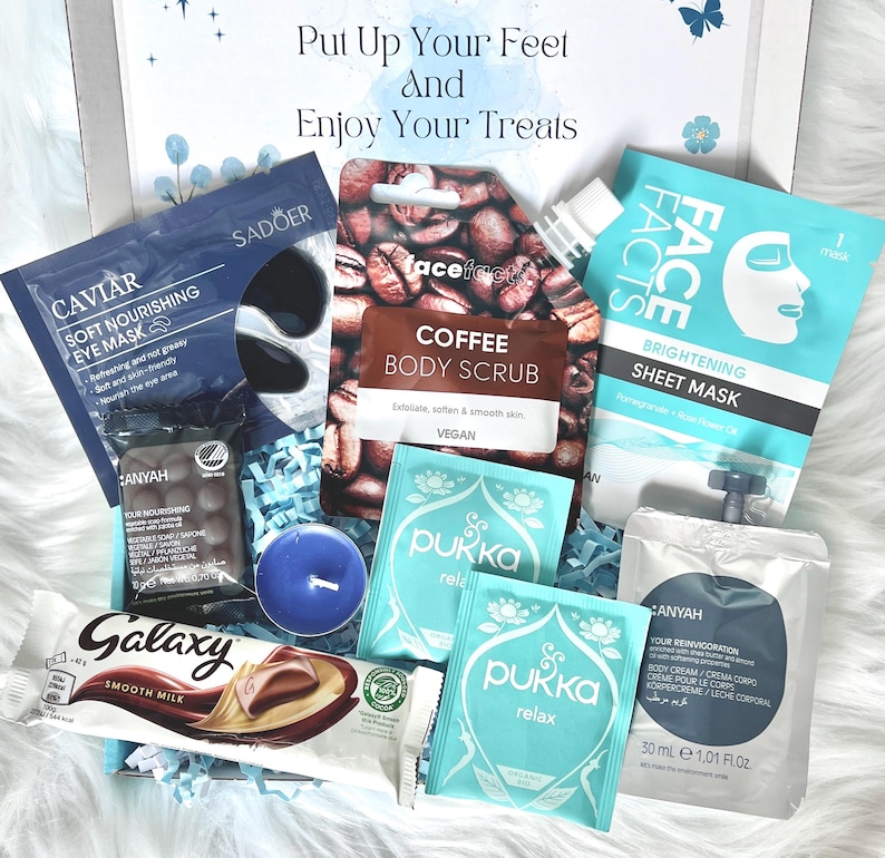 May include: A gift box filled with self-care items. Includes a coffee body scrub, eye mask, face sheet mask, chocolate bar, tea sachets, a candle, and body cream. The box has the text "Put Up Your Feet and Enjoy Your Treats".