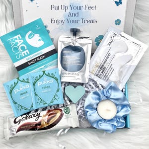 May include: A gift box filled with self-care items. Includes a face mask, eye mask, tea bags, a chocolate bar, a candle in a blue scrunchie, and a heart-shaped decoration. The box has the text "Put Up Your Feet and Enjoy Your Treats".
