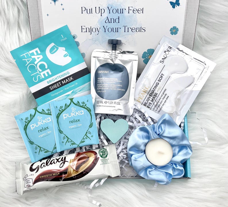 May include: A gift box filled with self-care items. Includes a face mask, eye mask, tea bags, a chocolate bar, a candle in a blue scrunchie, and a heart-shaped decoration. The box has the text "Put Up Your Feet and Enjoy Your Treats".