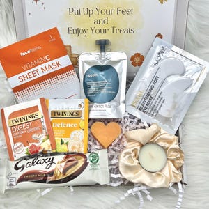 May include: A gift box filled with self-care items. Includes a Vitamin C sheet mask, tea bags, a chocolate bar, eye mask, a candle, and a heart-shaped soap. The box has text that reads "Put Up Your Feet and Enjoy Your Treats."