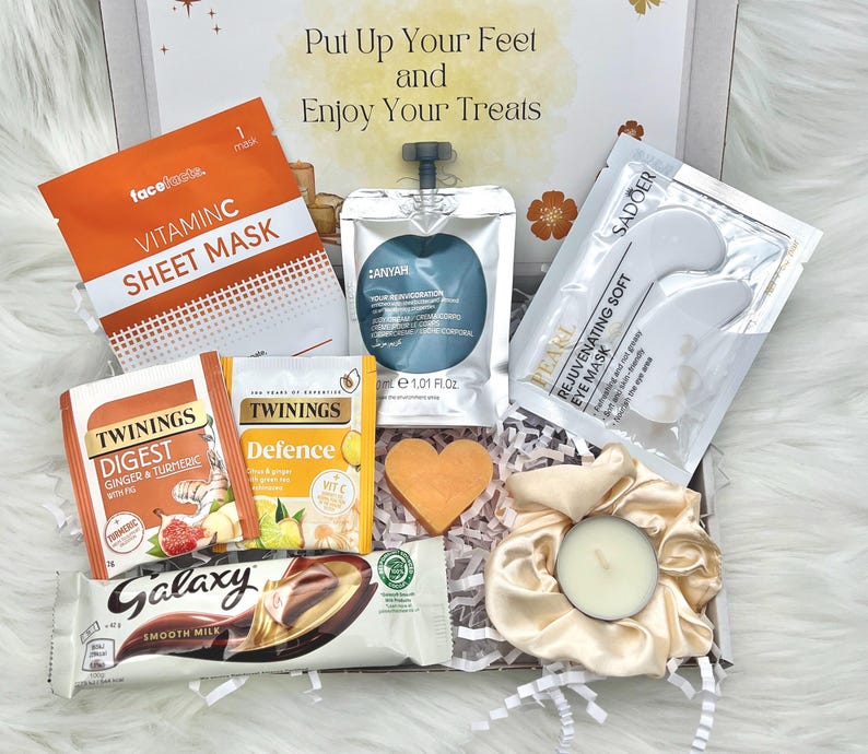 May include: A gift box filled with self-care items. Includes a Vitamin C sheet mask, tea bags, a chocolate bar, eye mask, a candle, and a heart-shaped soap. The box has text that reads "Put Up Your Feet and Enjoy Your Treats."
