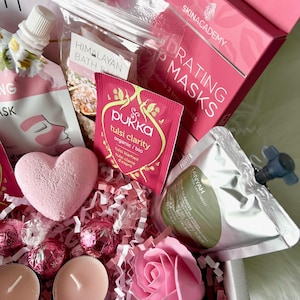 May include: A pink and white gift basket filled with bath and body products. The basket includes a pink bath bomb, a pink rose, two pink tea light candles, a small bag of Himalayan bath salts, a pouch of Tulsi Clarity tea, a pouch of face masks, and a bottle of body wash.
