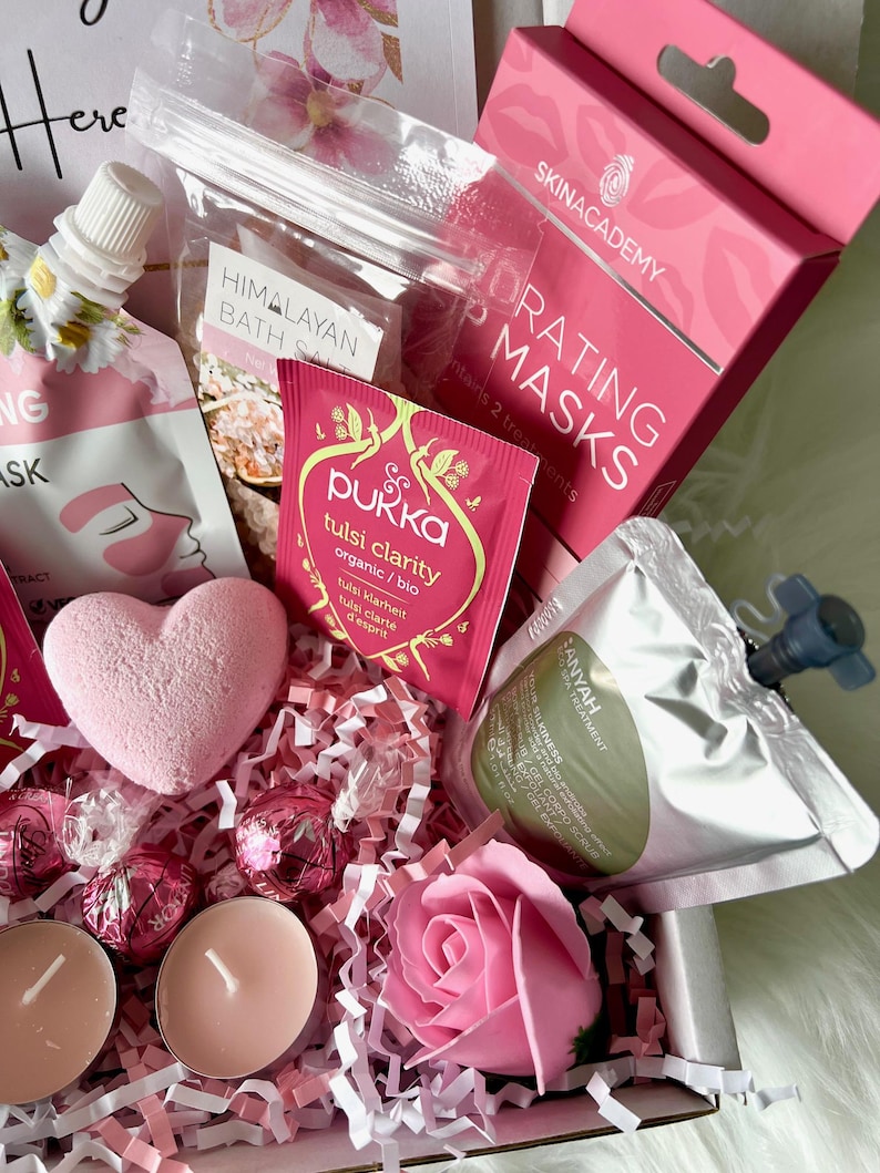 May include: A pink and white gift basket filled with bath and body products. The basket includes a pink bath bomb, a pink rose, two pink tea light candles, a small bag of Himalayan bath salts, a pouch of Tulsi Clarity tea, a pouch of face masks, and a bottle of body wash.