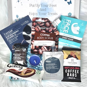 May include: A gift box filled with self-care products. Includes a coffee body scrub, eye mask, sheet mask, body cream, coffee bags, a Galaxy chocolate bar, and a blue candle. The box has the text "Put Up Your Feet And Enjoy Your Treats".
