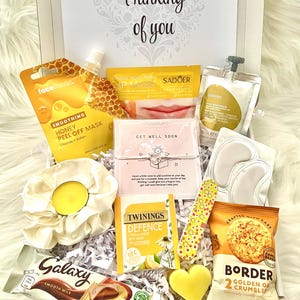 May include: A gift box filled with self-care items, including a face mask, lip mask, eye patches, tea, chocolate bar, and snacks. The box is decorated with the phrase "Thinking of you" and includes a scrunchie and a heart-shaped soap.